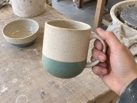 First-person phone snapshot: the client holds the cream-and-teal ceramic mug by its handle above the studio table