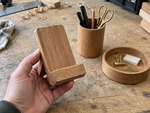 First-person phone snapshot: the client holds a wooden phone stand above the workshop table, with the matching pen holder and tray behind it
